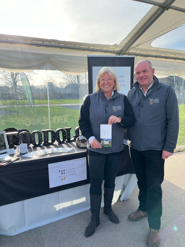 Two smiling vendors stand in front of a display table presenting luxury toilet hire services 