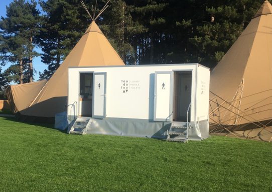 Two portable toilets next to large tan tipis on a grassy field.