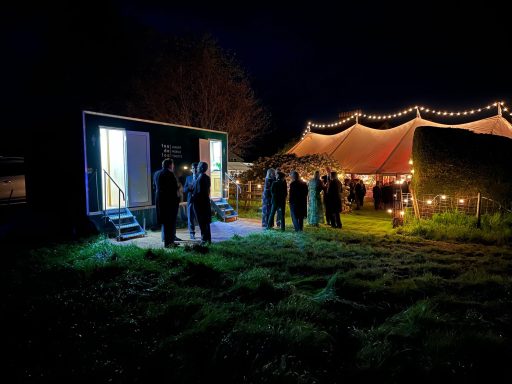 People gathered outside a tent at night, with a lit mobile unit nearby.