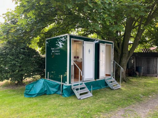 Two green portable bathroom units with steps, surrounded by trees in a grassy area.