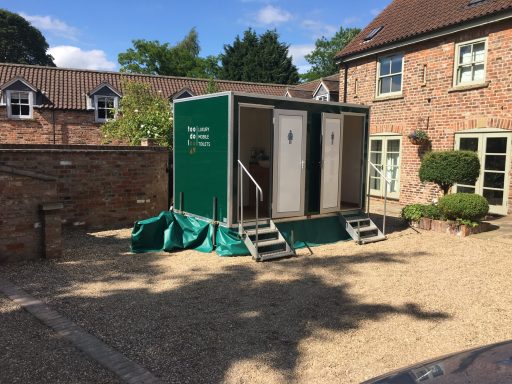 Green portable toilets on a wooden platform in a garden area with gravel and surrounding buildings.