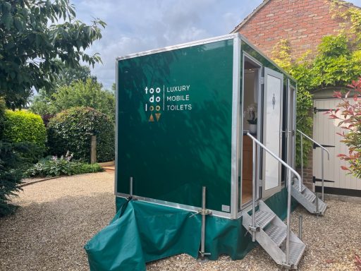 Green portable building with steps, set in a garden area with trees and shrubs.