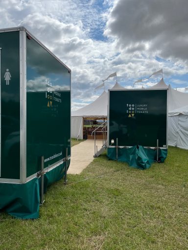 Green portable toilets beside a pathway leading to a marquee under a cloudy sky.